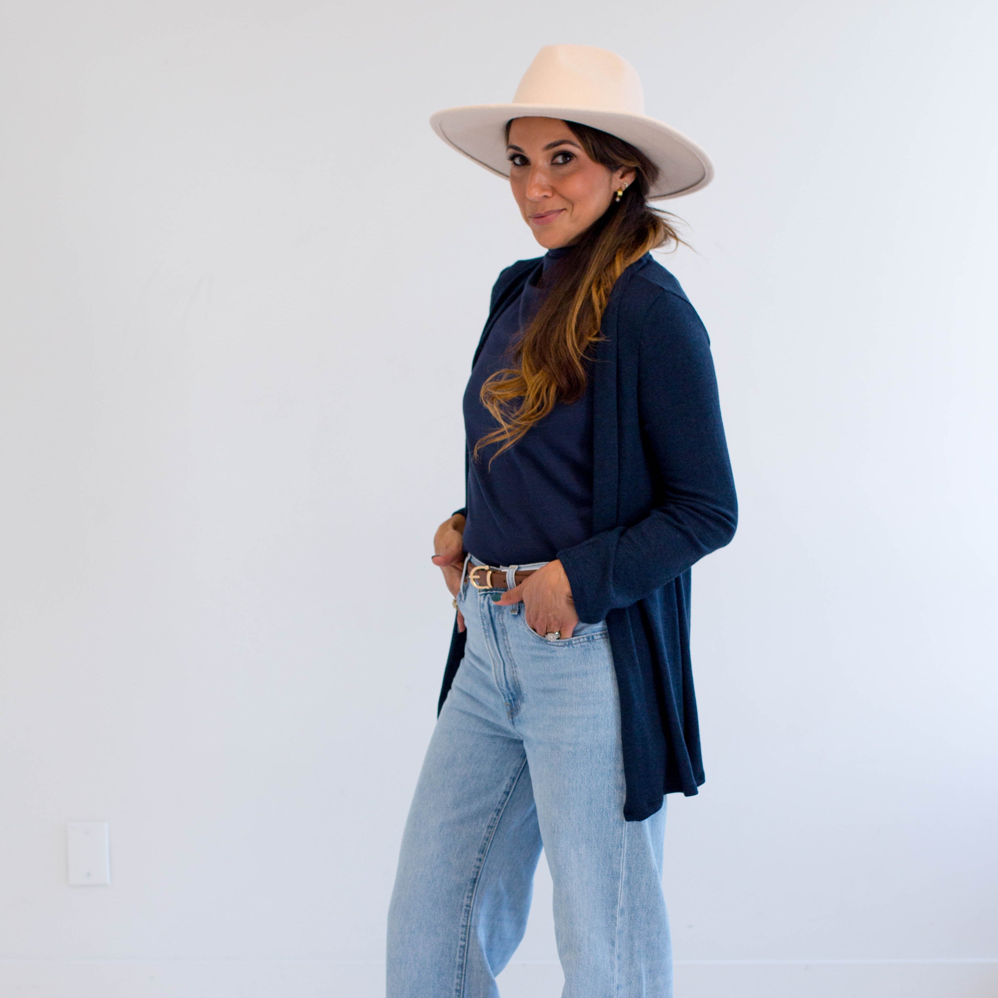 Woman wearing a navy cardigan made in canada, light blue jeans, and a beige hat on a white background