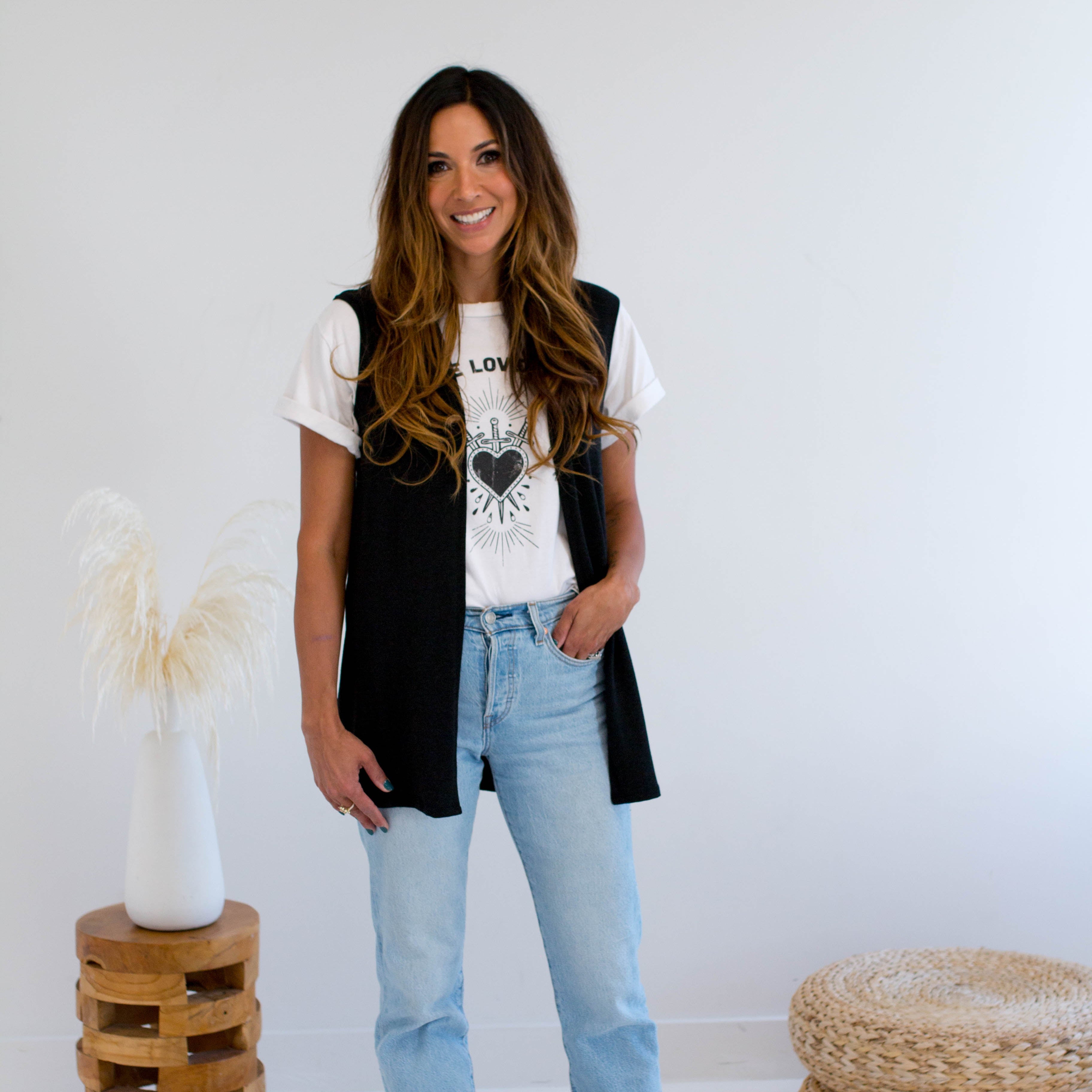 Woman wearing a black vest made in canada, white t-shirt with a heart design, and light blue jeans standing in a minimalistic room.
