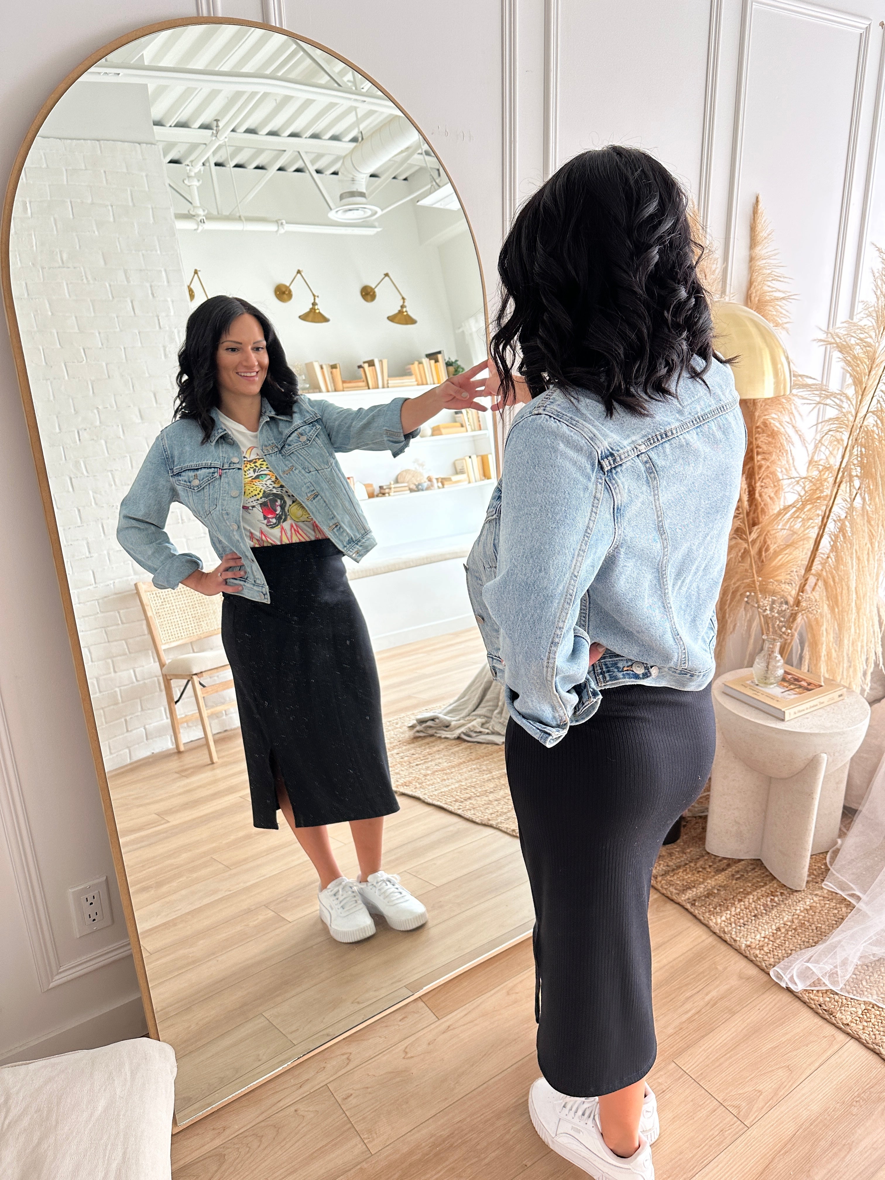 woman wearing denim jacket and black ribbed midi skirt with white sneakers standing looking in an arched mirror