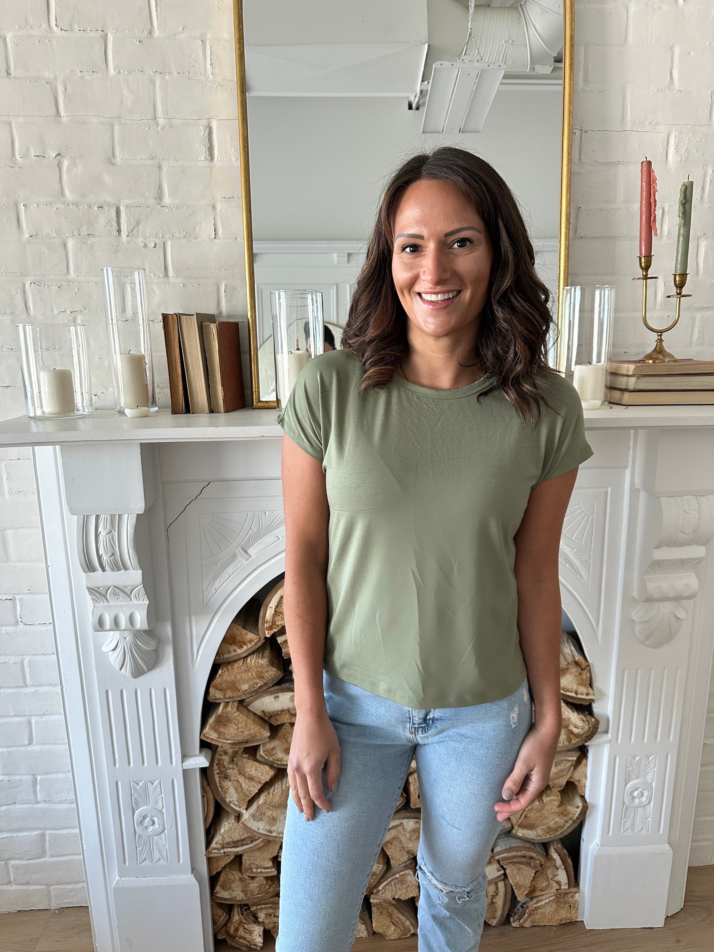 woman smiling standing wearing a sage green bamboo t-shirt and jeans. Standing in front of a fireplace.