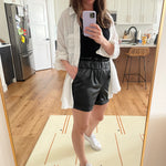 woman taking a selfie wearing a white button up blouse and black leather paperbag shorts