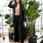 woman wearing a leather blazer, white graphic tee and black wide leg pants and pointy toed shoes next to a plant