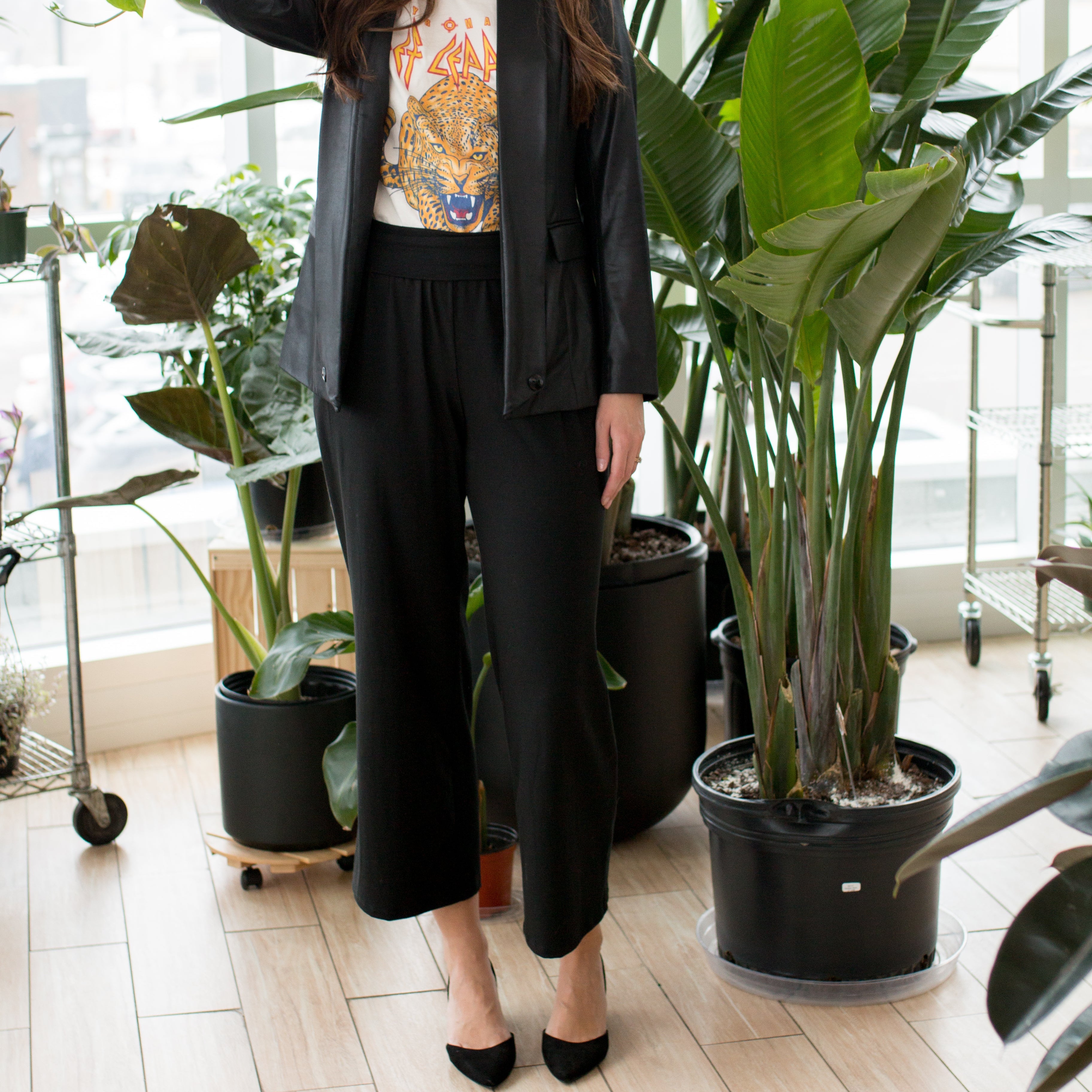 woman wearing a leather blazer, white graphic tee and black wide leg pants and pointy toed shoes next to a plant