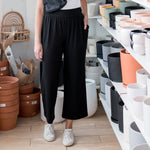 Brunette woman wearing a white t shirt and black wide leg pants standing in a room with plant pots