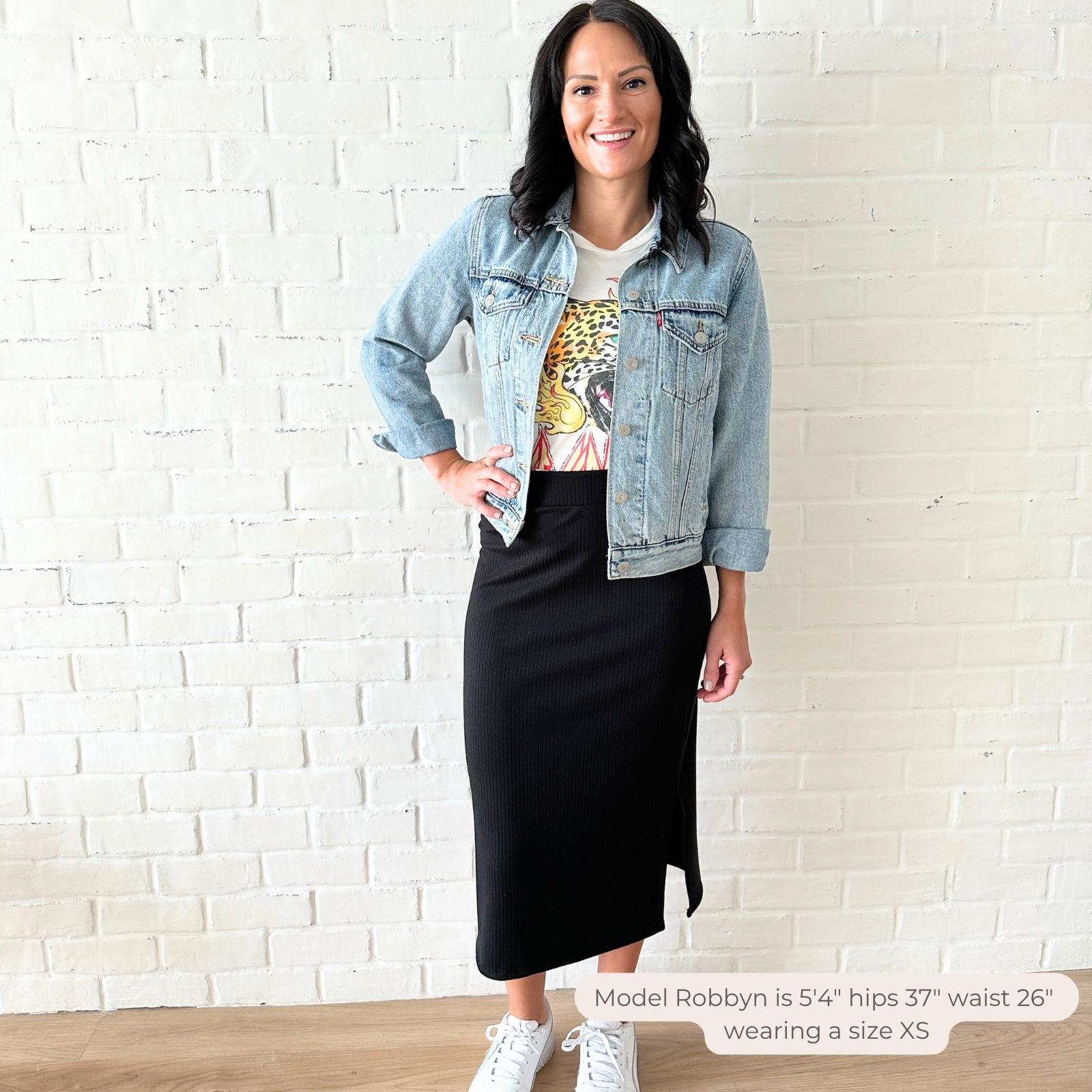 Woman wearing a denim jacket, graphic t-shirt, and black midi skirt against a white brick wall. Apparel made in canada by ang hill
