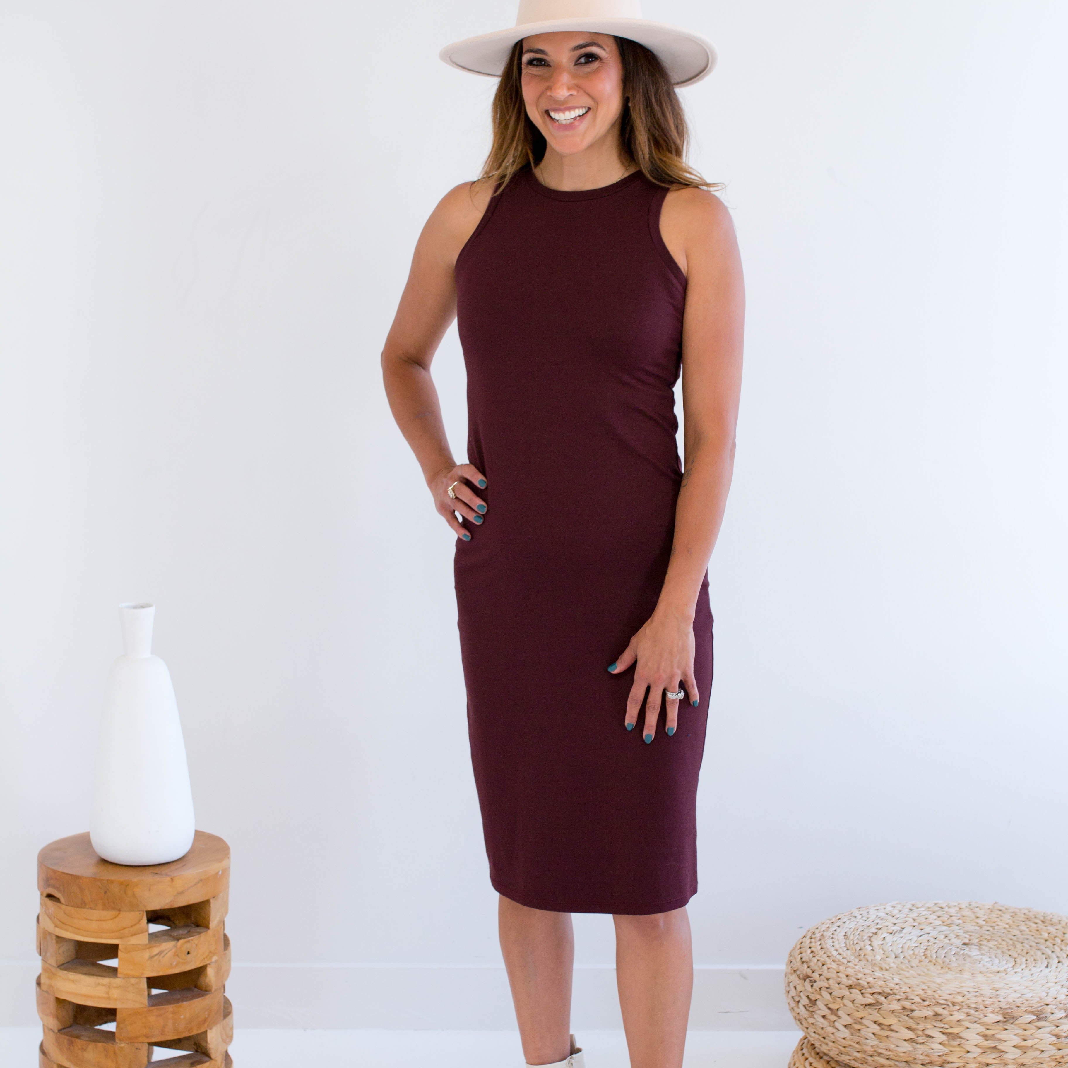 Woman wearing a burgundy tank dress made in canada with a white hat and boots, standing against a white background.