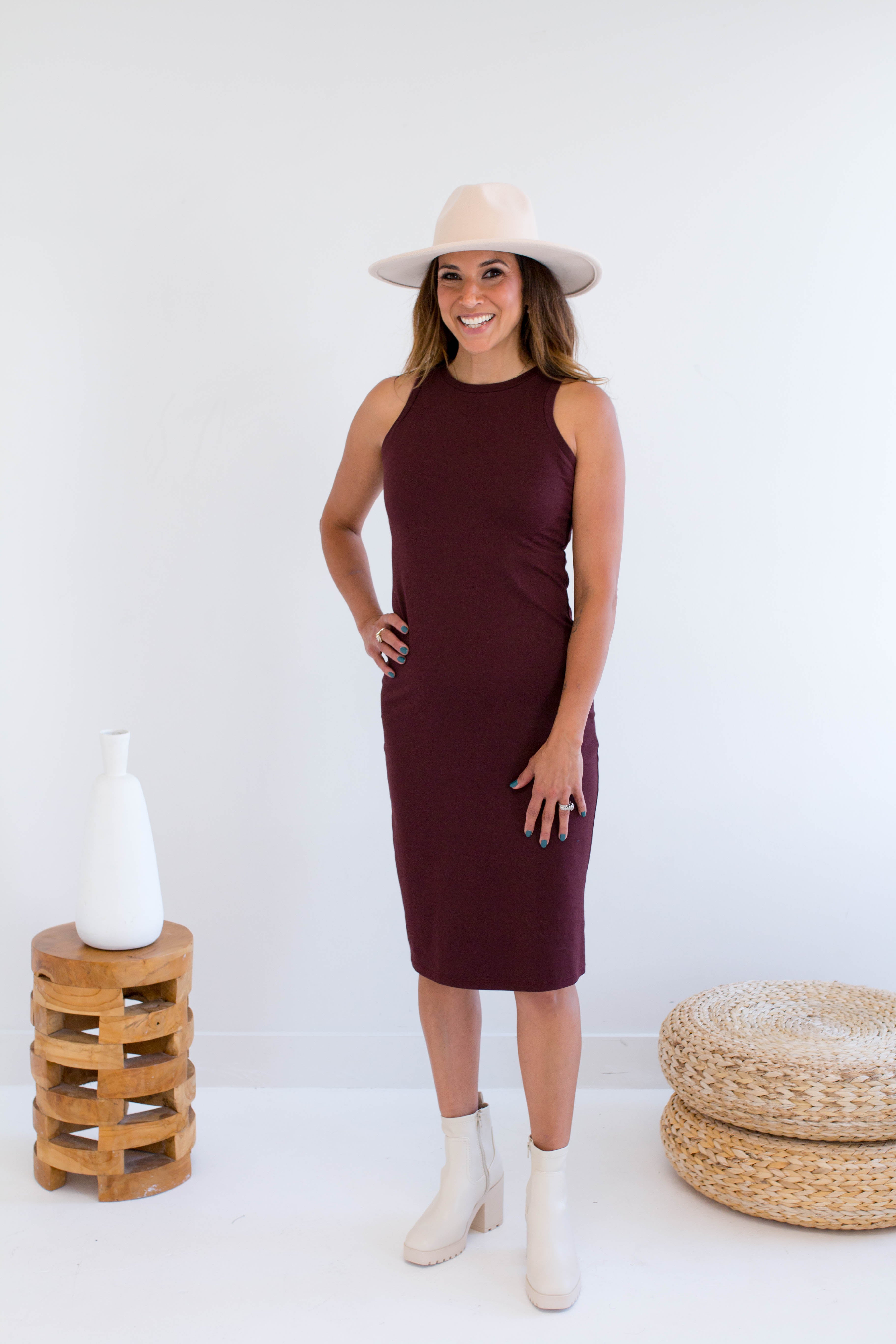Woman wearing a burgundy tank dress made in canada with a white hat and boots, standing against a white background.