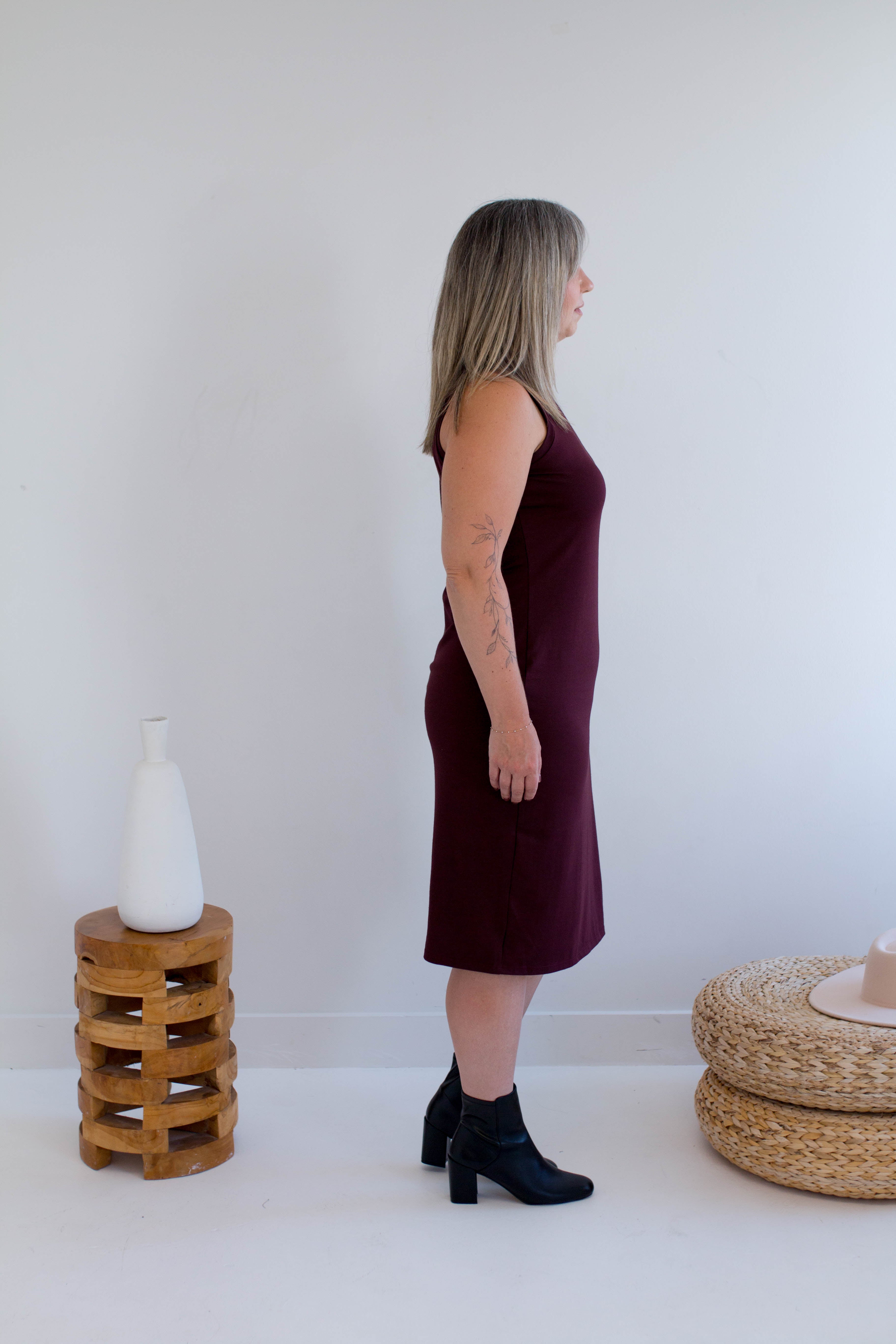 Woman wearing a burgundy tank dress made in canada standing in a minimalistic room with a white wall and wooden furniture.