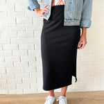 Person wearing a black ribbed skirt with a denim jacket and white sneakers against a white brick wall.