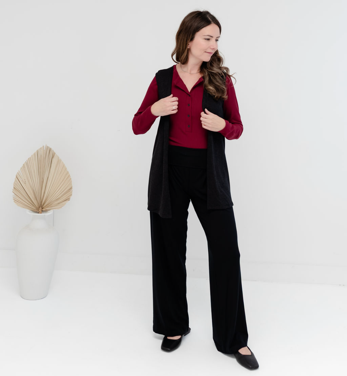A woman modeling a black sleeveless vest over a burgundy long-sleeve blouse and black wide-leg trousers, standing in a bright white studio with a decorative dried palm in a white vase.