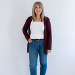 Woman wearing a burgundy cardigan made in canada, white shirt, blue jeans, and black boots against a white background