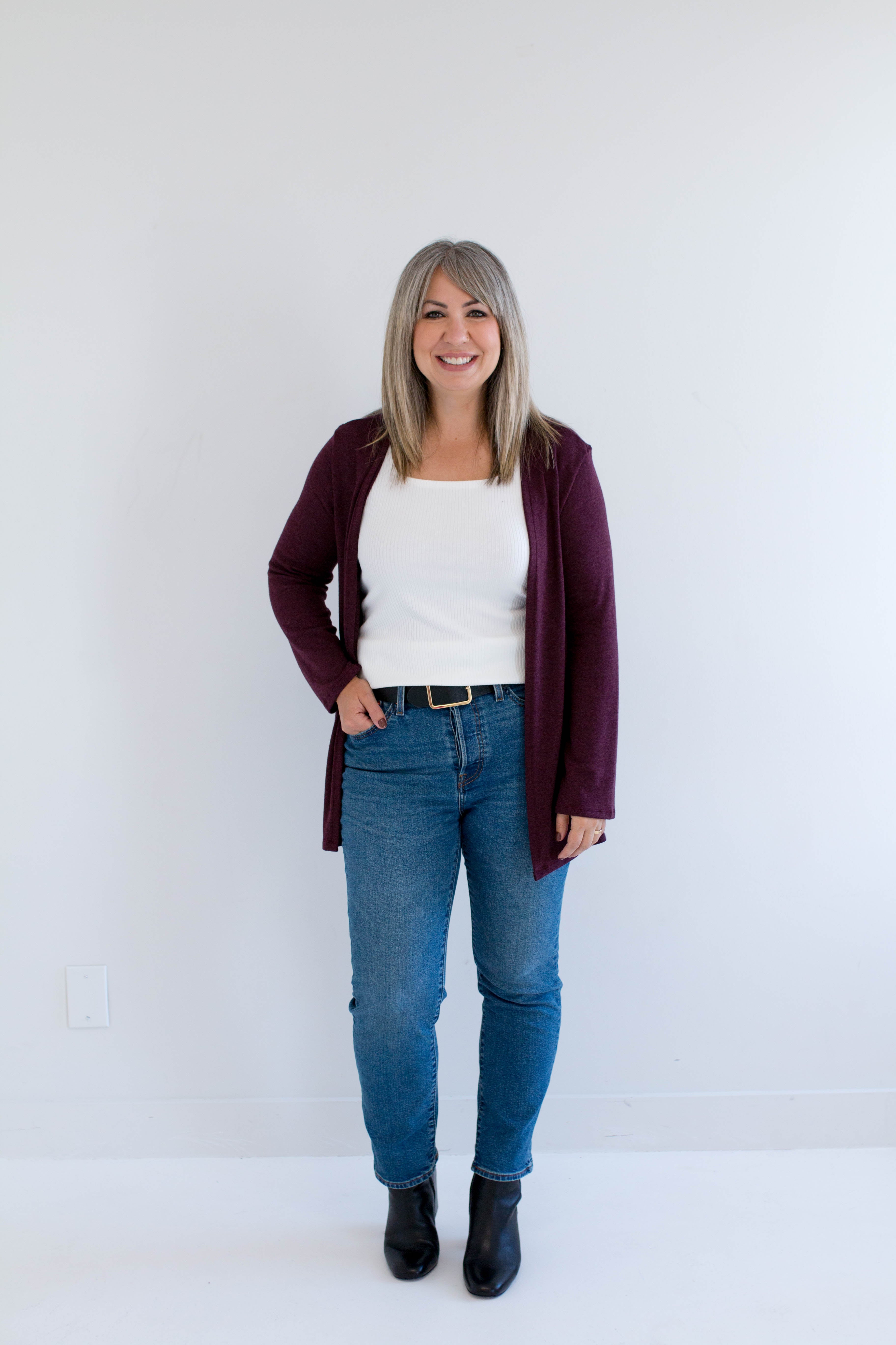 Woman wearing a burgundy cardigan made in canada, white shirt, blue jeans, and black boots against a white background