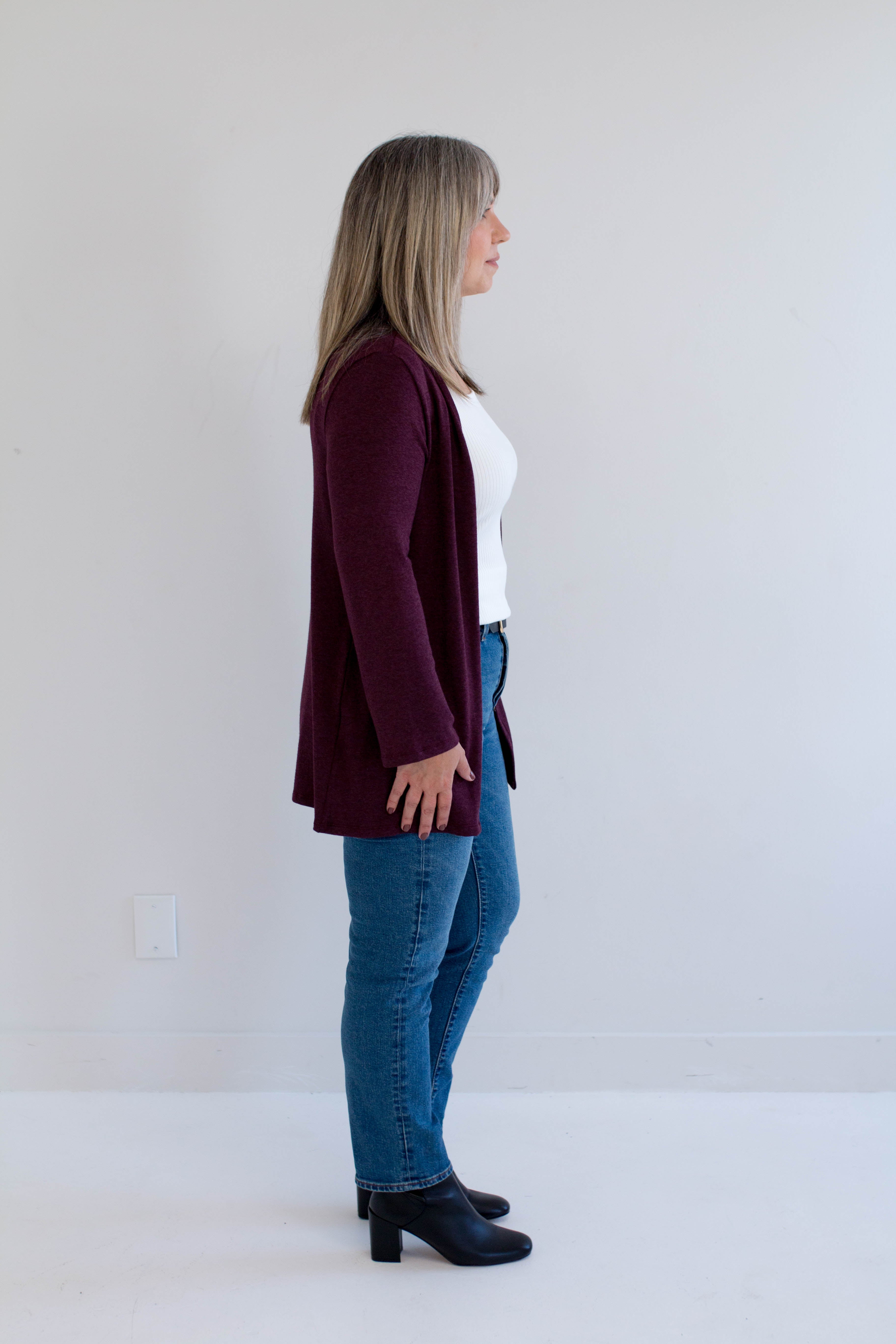 Woman wearing a burgundy cardigan, white shirt, blue jeans, and black boots against a white background