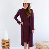Woman wearing a long merlot dress and long burgundy cardigan with a matching hat and white boots, standing against a white background.