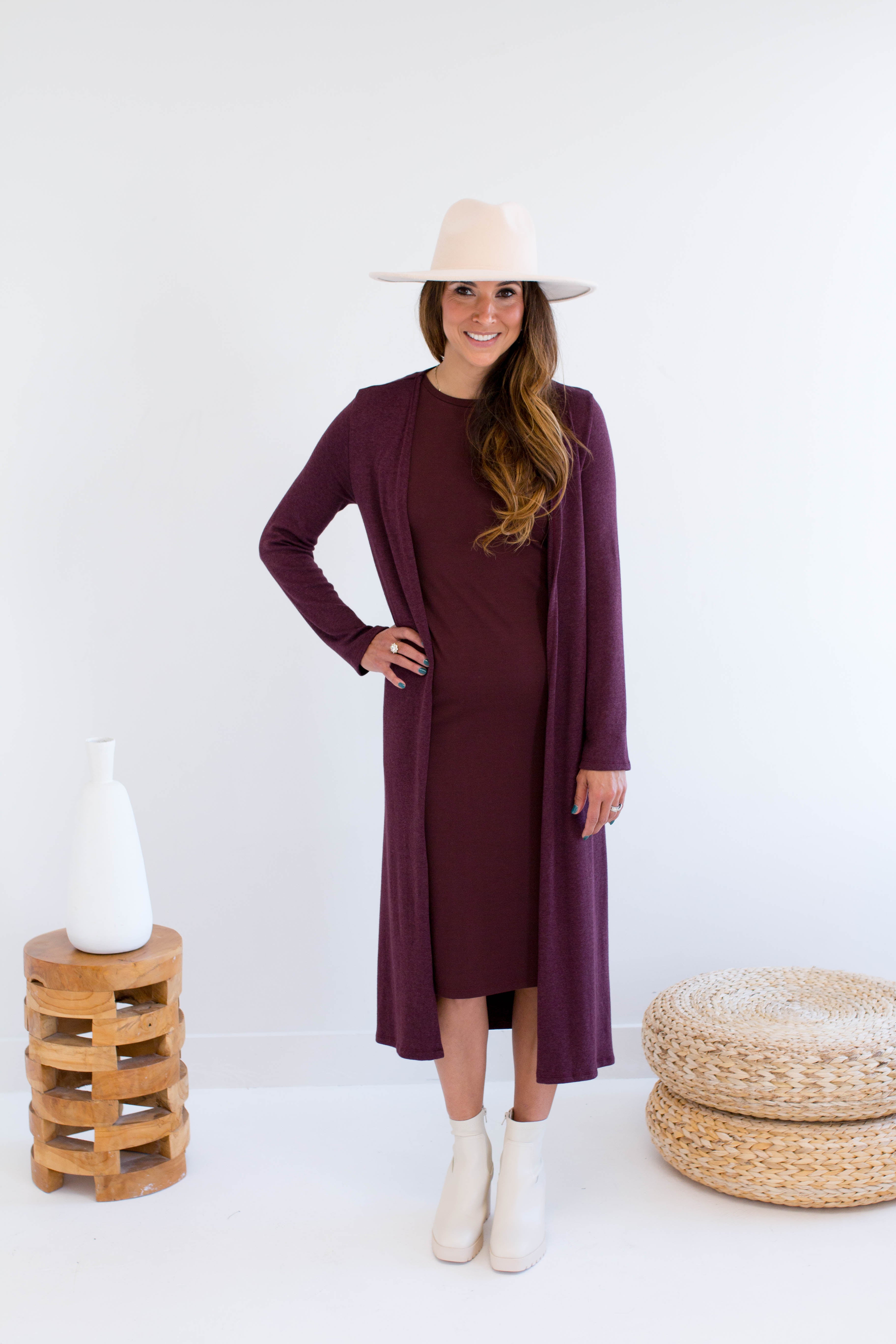 Woman wearing a long merlot dress and long burgundy cardigan with a matching hat and white boots, standing against a white background.