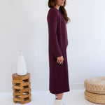 Woman wearing a long burgundy tank dress and merlot long duster cardigan,  white boots, standing in a minimalistic room with a white wall and wooden stool.