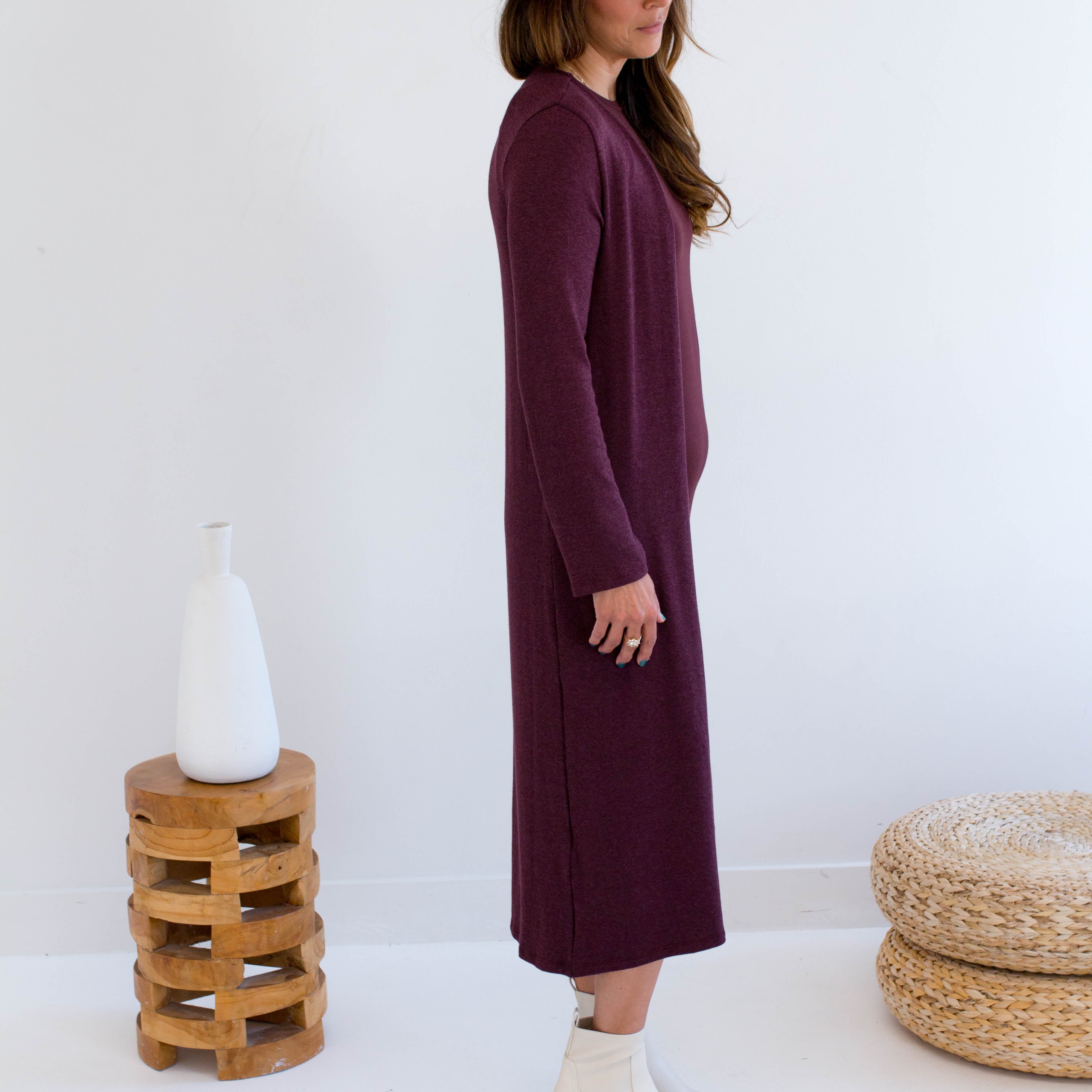 Woman wearing a long burgundy tank dress and merlot long duster cardigan,  white boots, standing in a minimalistic room with a white wall and wooden stool.