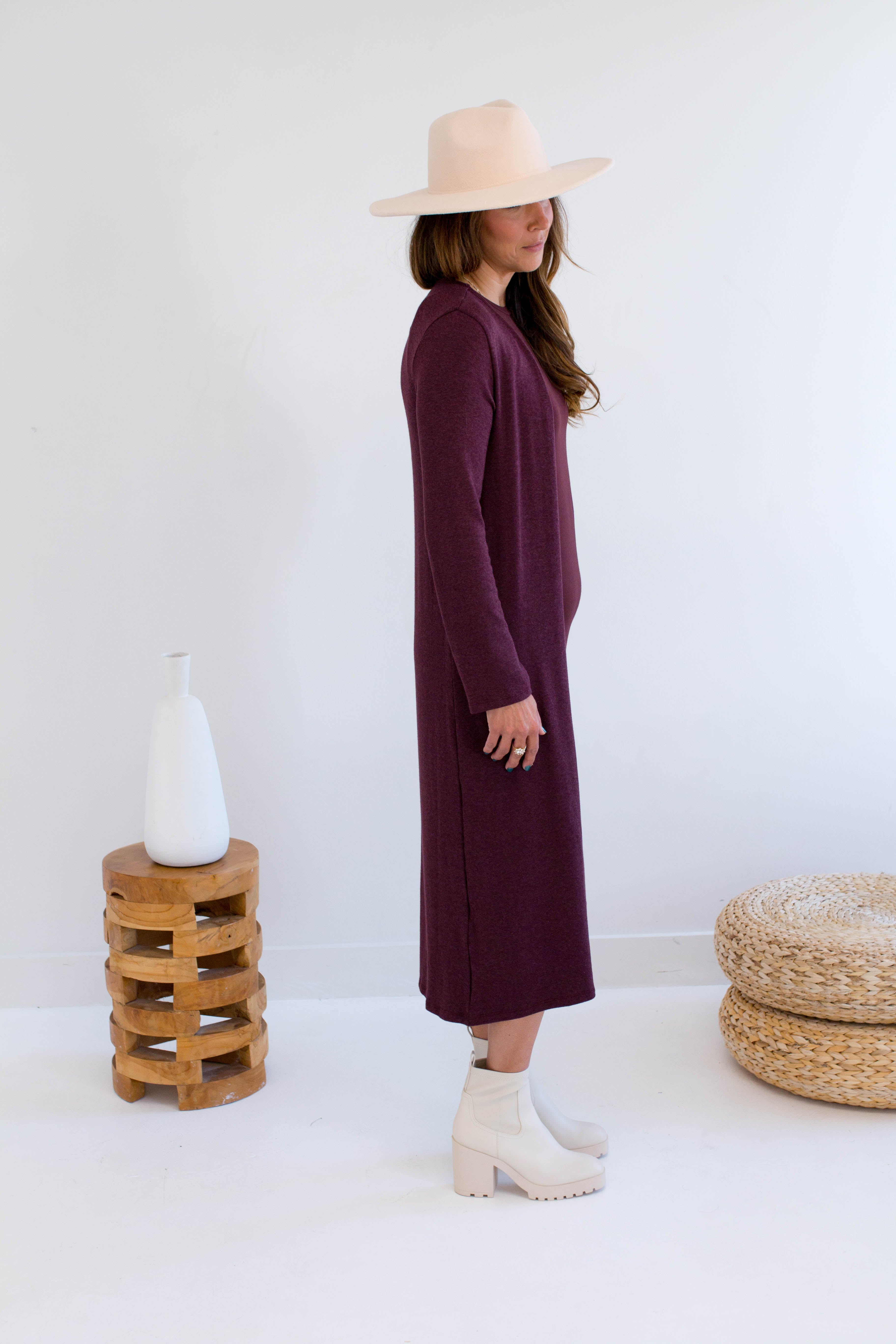 Woman wearing a long burgundy tank dress and merlot long duster cardigan,  white boots, standing in a minimalistic room with a white wall and wooden stool.