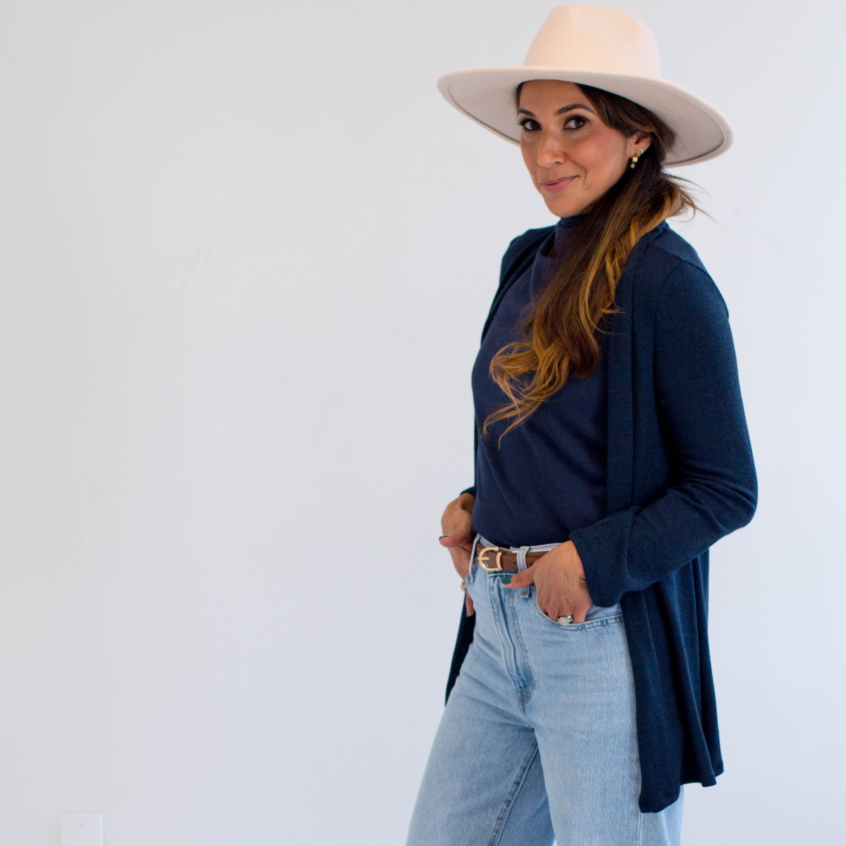 Woman wearing a navy cardigan made in canada, light blue jeans, and a beige hat on a white background