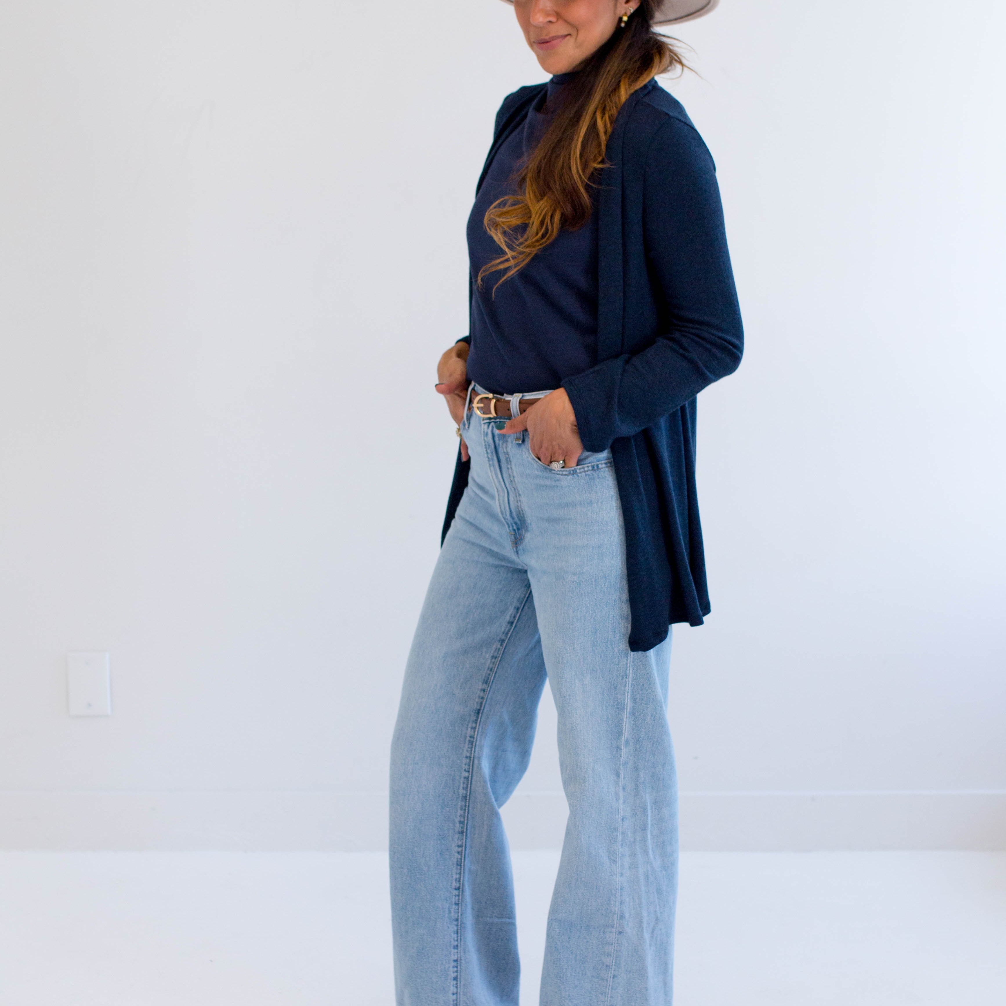 Woman wearing a navy cardigan made in canada, light blue jeans, and a beige hat on a white background