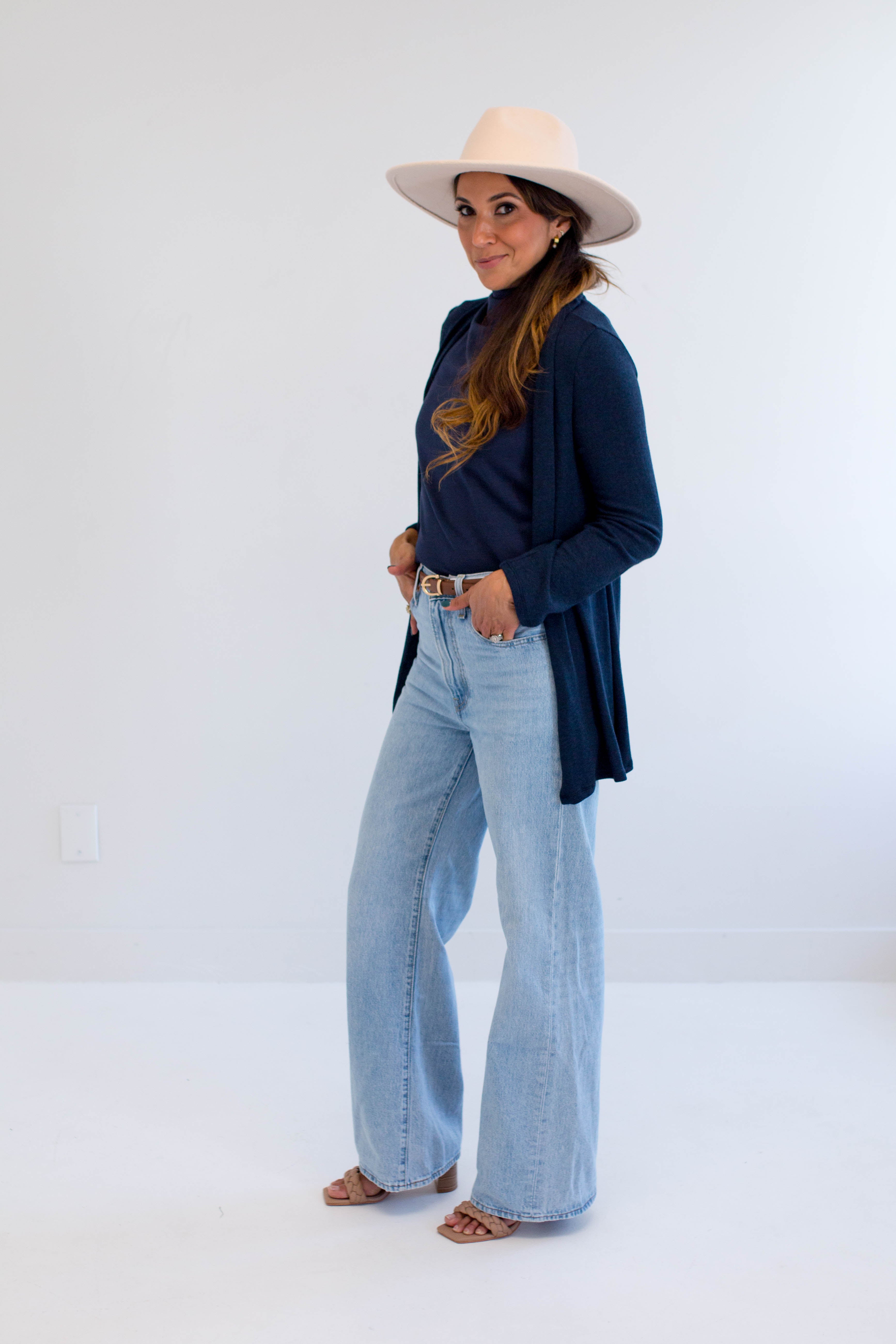 Woman wearing a navy cardigan made in canada, light blue jeans, and a beige hat on a white background
