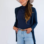 Woman wearing a navy cardigan made in canada, navy mockneck, light blue jeans, and a beige hat against a white background
