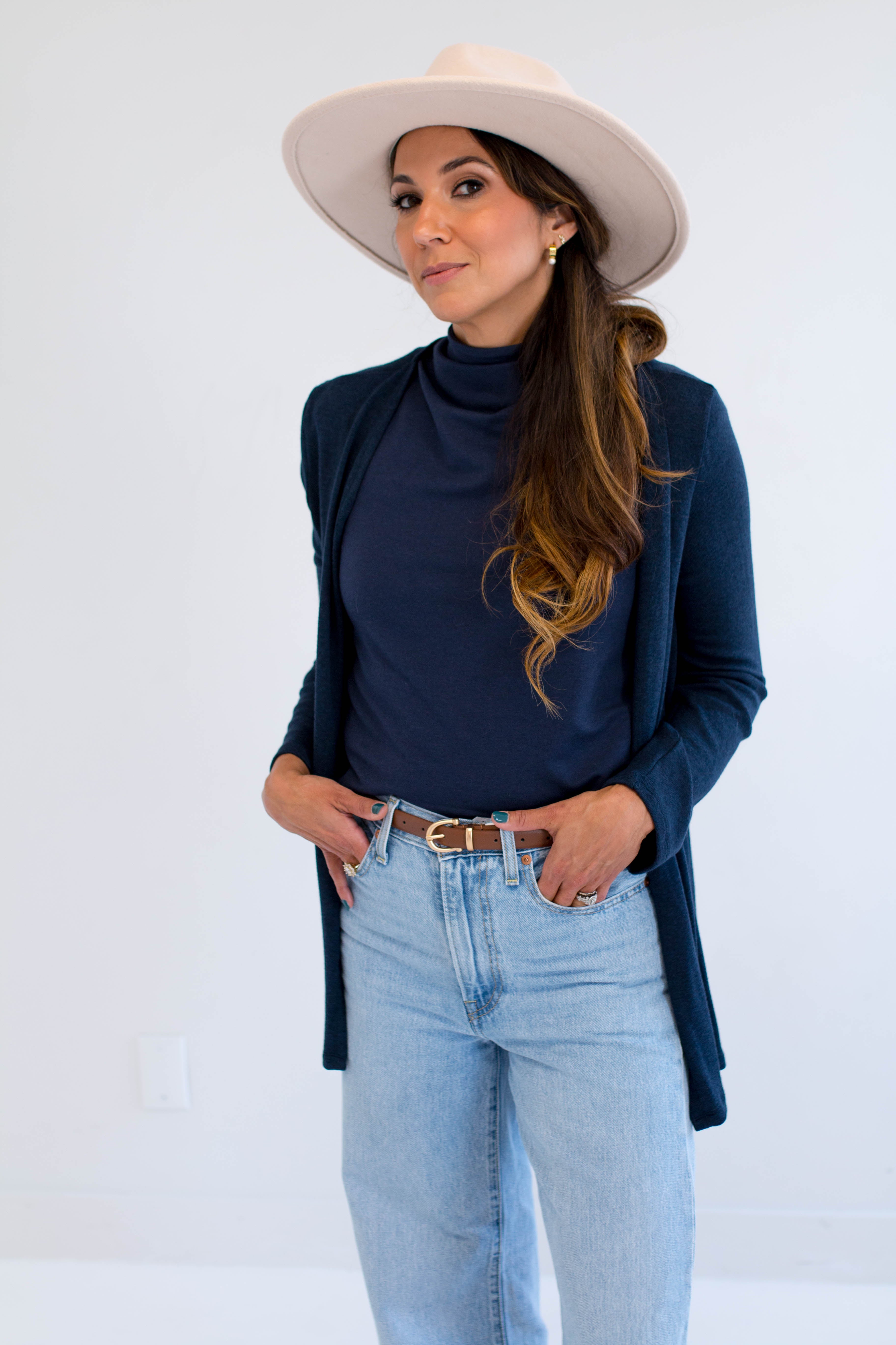 Woman wearing a navy cardigan made in canada, navy mockneck, light blue jeans, and a beige hat against a white background