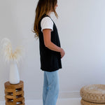 Woman wearing a black vest made in canada and light blue jeans standing in a minimalistic room with a white wall and wooden stool.