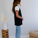 Woman wearing a black vest made in canada and light blue jeans standing in a minimalistic room with a white wall and wooden stool.