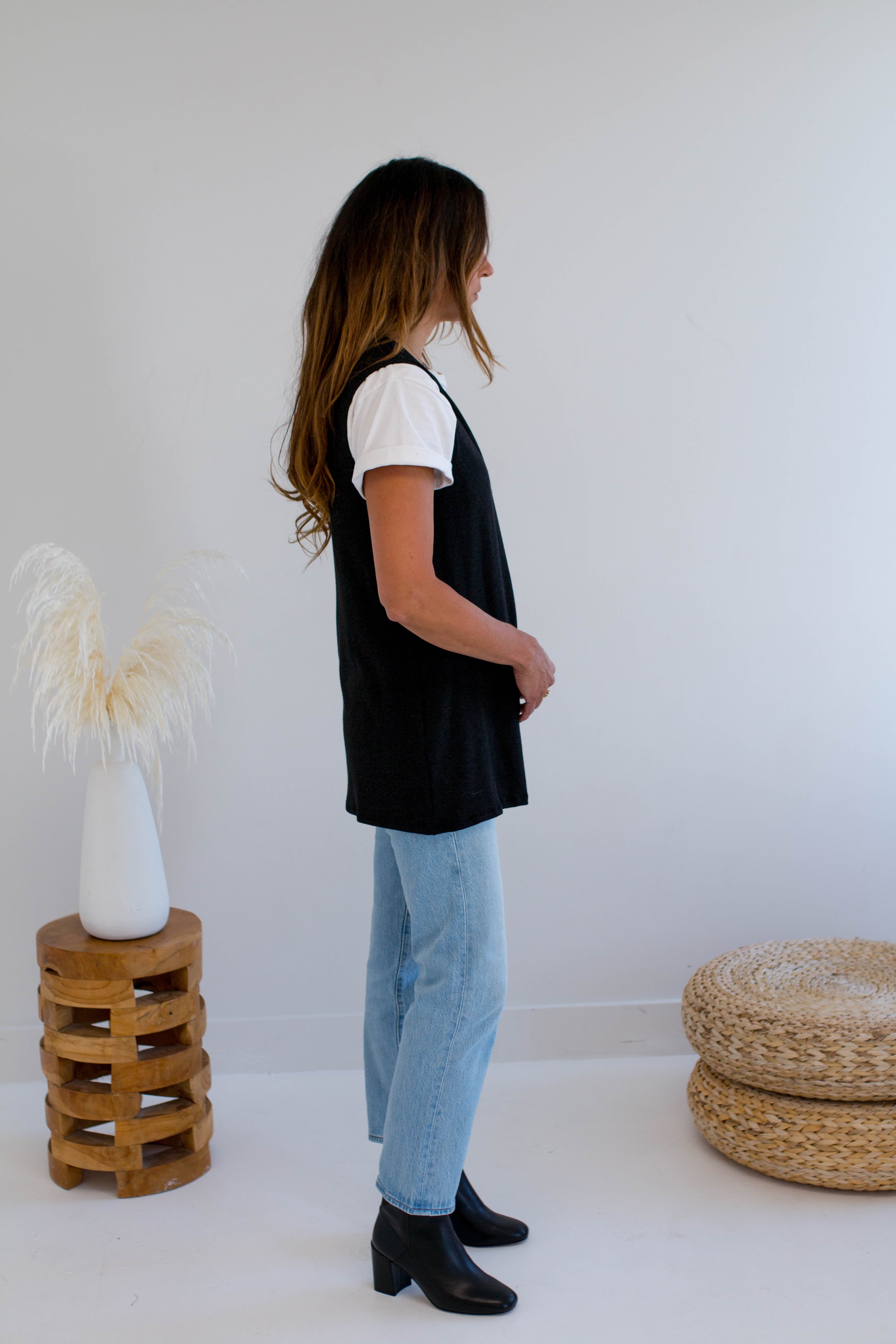 Woman wearing a black vest made in canada and light blue jeans standing in a minimalistic room with a white wall and wooden stool.