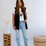 Woman wearing a black vest made in canada, white t-shirt with a heart design, and light blue jeans standing in a minimalistic room.