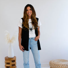 Woman wearing a black vest made in canada, white t-shirt with a heart design, and light blue jeans standing in a minimalistic room.