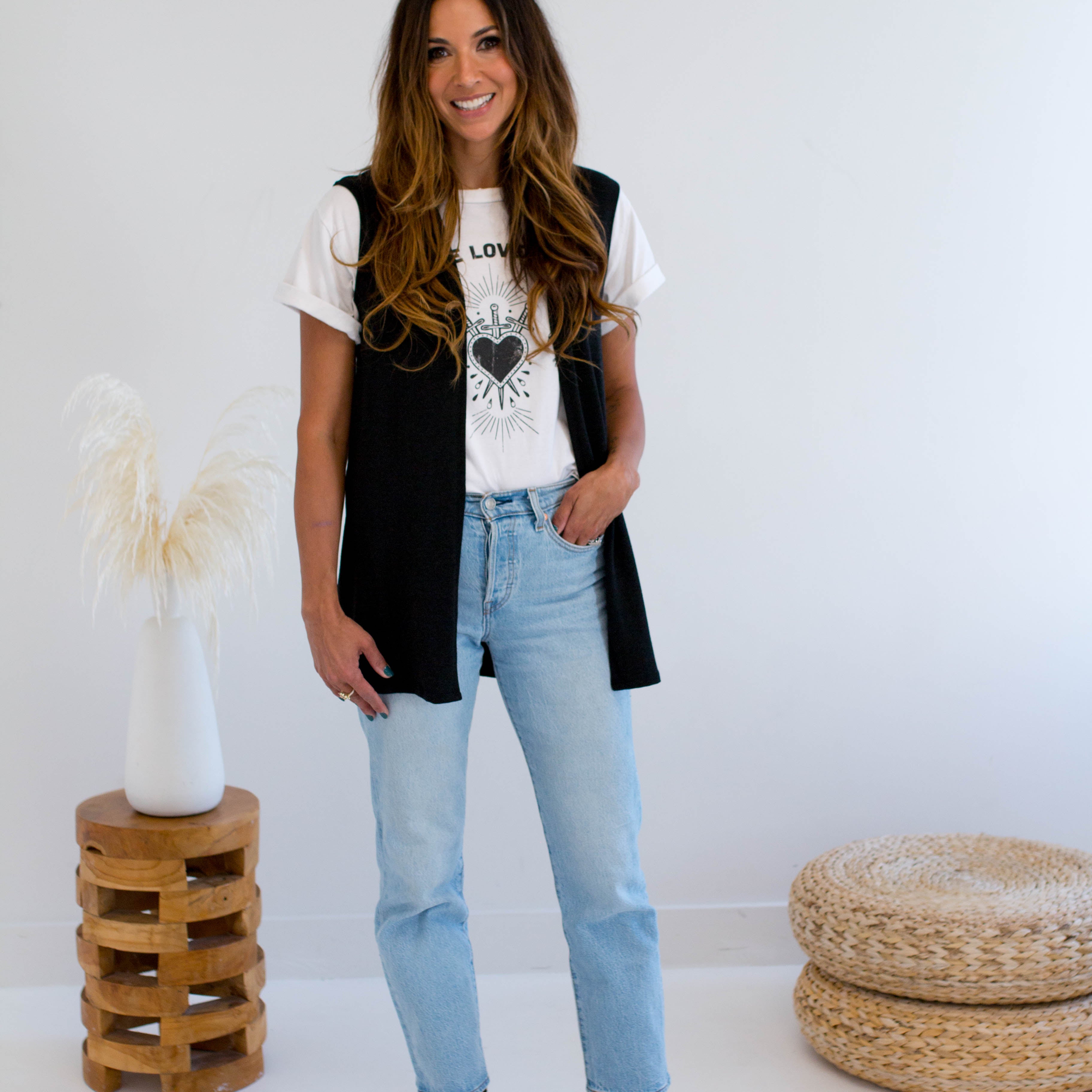 Woman wearing a black vest made in canada, white t-shirt with a heart design, and light blue jeans standing in a minimalistic room.