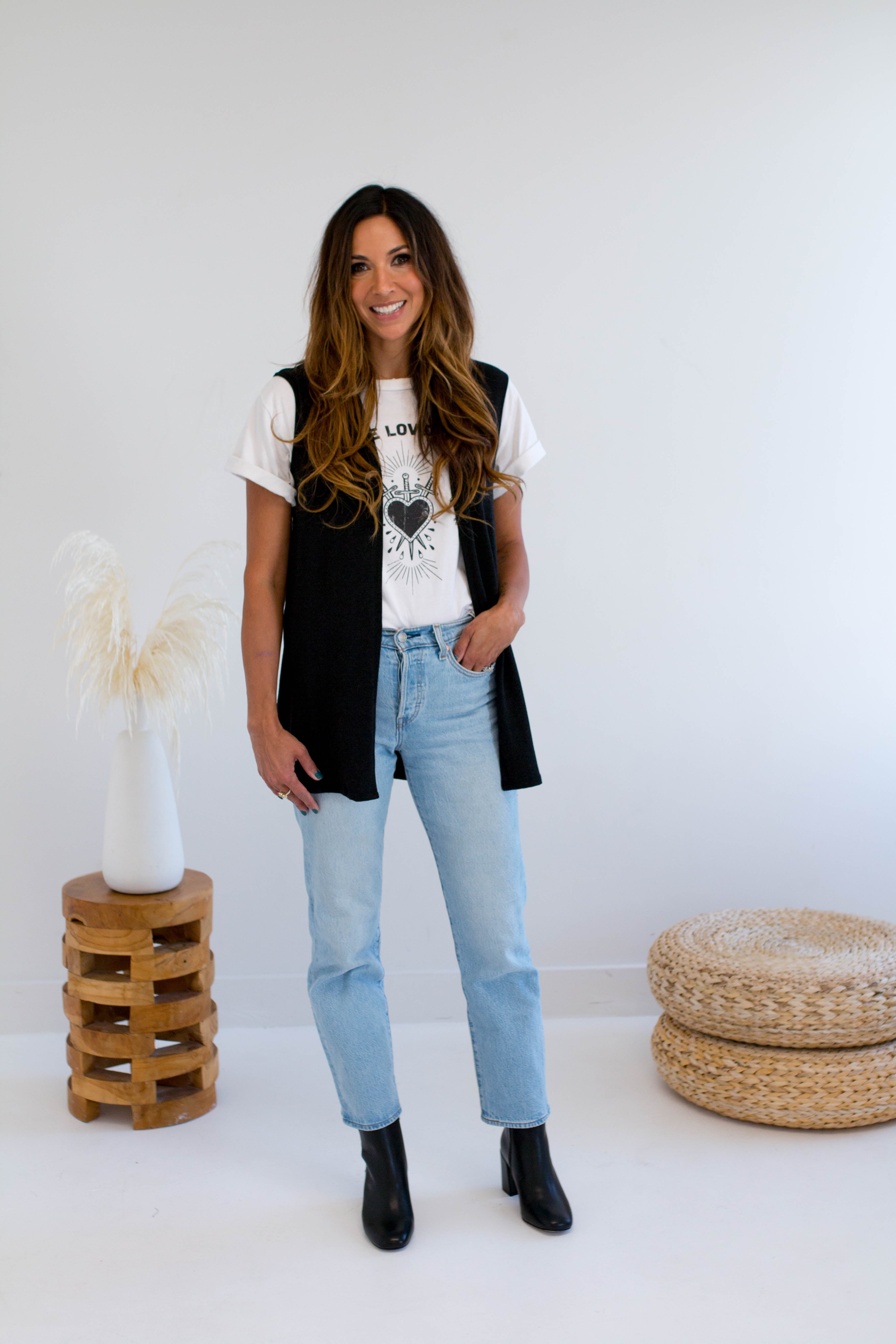 Woman wearing a black vest made in canada, white t-shirt with a heart design, and light blue jeans standing in a minimalistic room.