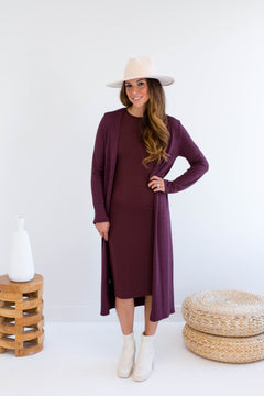 Woman wearing a long burgundy cardigan over a dress made in canada, standing in a minimalistic setting with a white background.