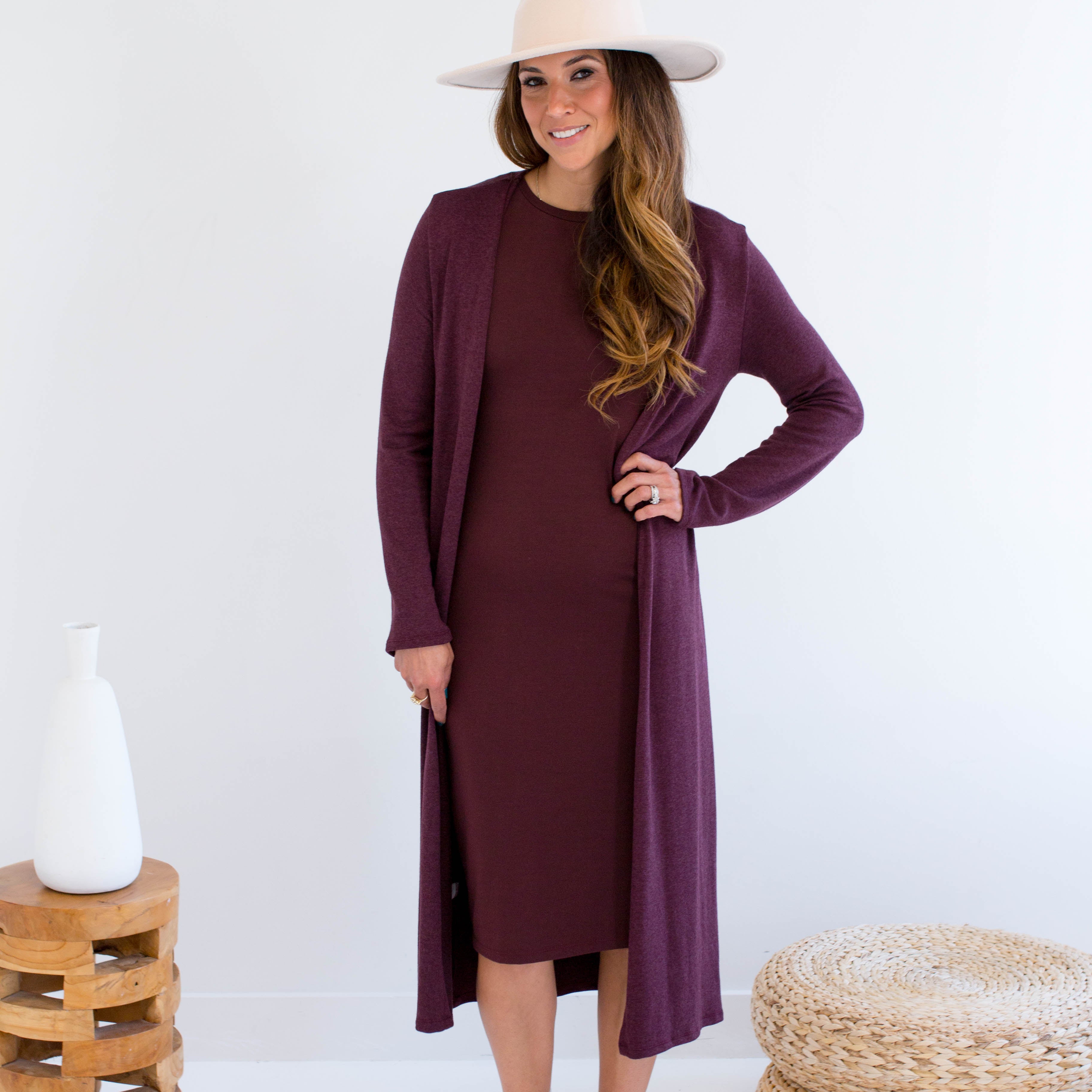 Woman wearing a long burgundy cardigan over a dress made in canada, standing in a minimalistic setting with a white background.
