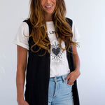 Woman wearing a black sleeveless jacket over a white t-shirt with a graphic design, light blue jeans, and a white background. Made in Canada.