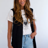 Woman wearing a black sleeveless jacket over a white t-shirt with a graphic design, light blue jeans, and a white background. Made in Canada.