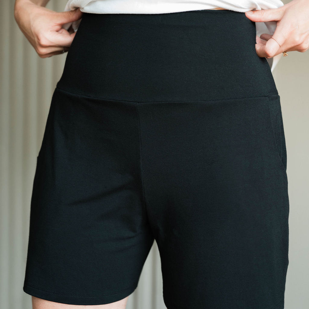 A close-up of a person wearing black high-waisted athletic shorts with a white top, showing the fit and side pockets. Made in Canada bamboo shorts.