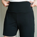 A close-up of a person wearing black high-waisted athletic shorts with a white top, showing the fit and side pockets. Made in Canada bamboo shorts.