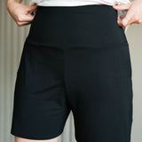 A close-up of a person wearing black high-waisted athletic shorts with a white top, showing the fit and side pockets. Made in Canada bamboo shorts.