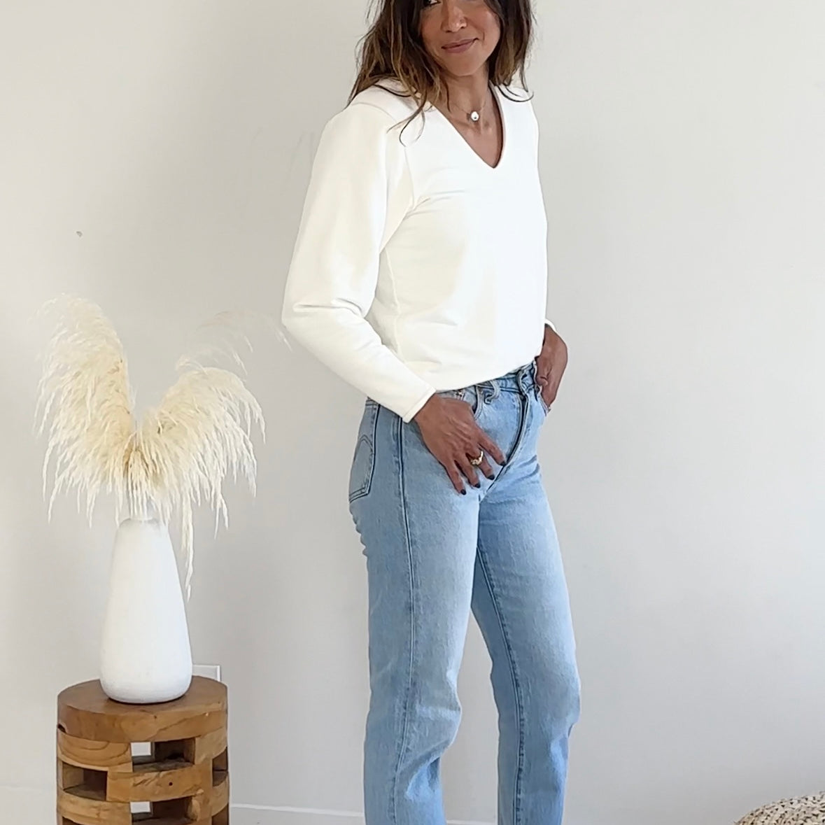 Woman wearing a off-white sweater and light blue jeans standing in a minimalistic room.