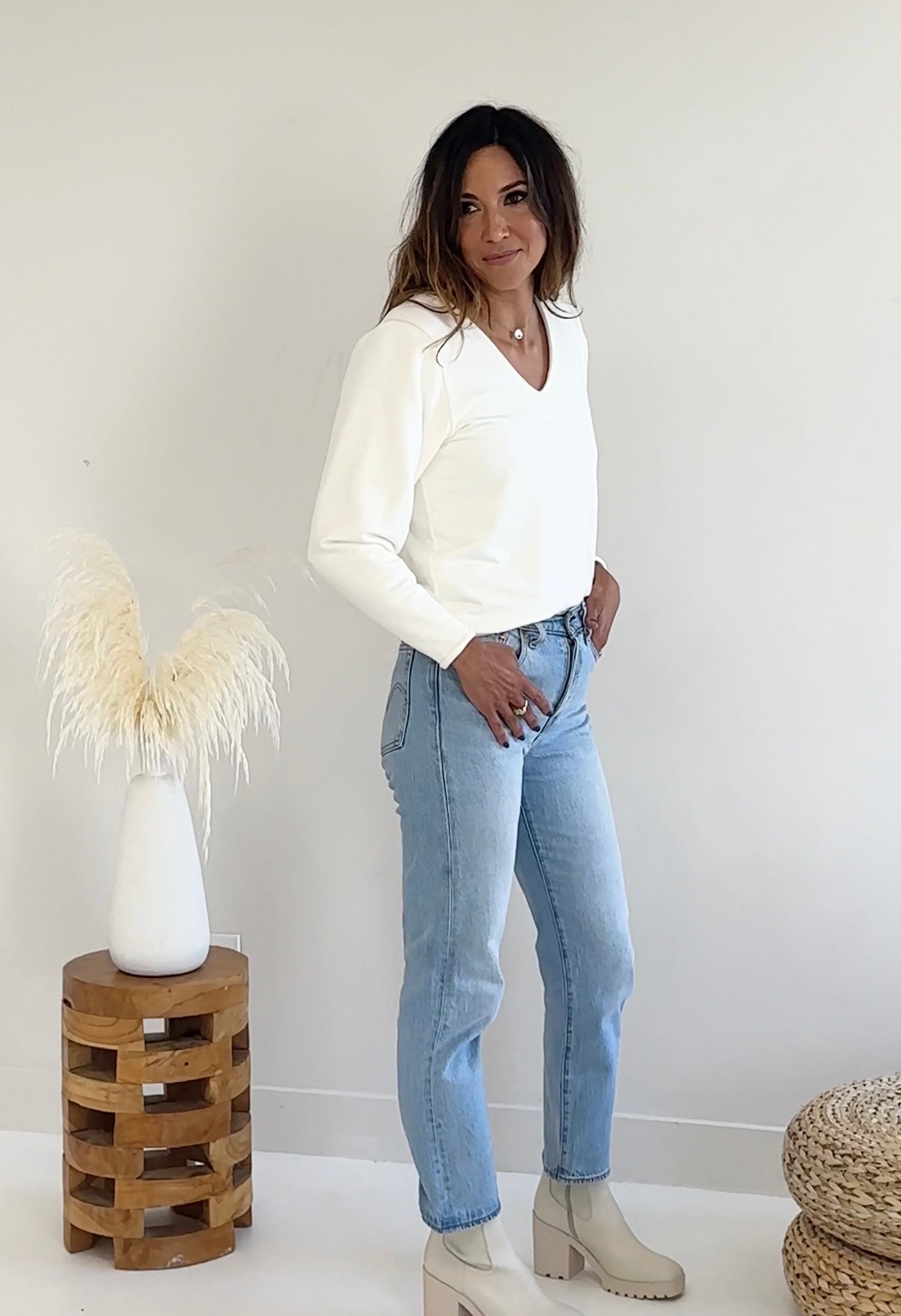 Woman wearing a off-white sweater and light blue jeans standing in a minimalistic room.