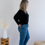 Woman wearing a black v-neck sweater and blue jeans standing in a minimalistic room with a white wall and wooden stool.