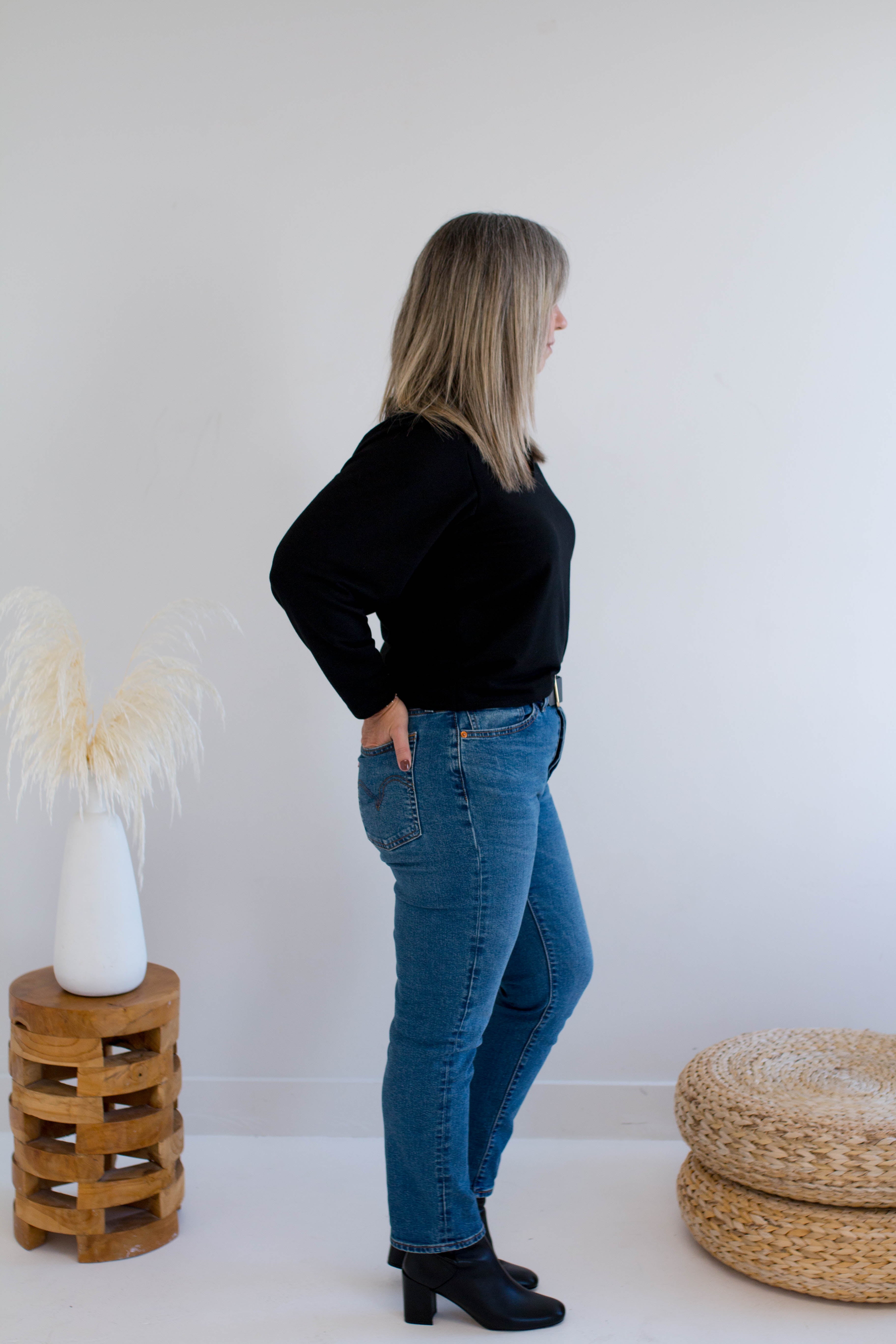 Woman wearing a black v-neck sweater and blue jeans standing in a minimalistic room with a white wall and wooden stool.