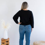 back view of a woman wearing a black v-neck sweater and blue jeans standing against a white wall with decorative items.