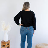 back view of a woman wearing a black v-neck sweater and blue jeans standing against a white wall with decorative items.