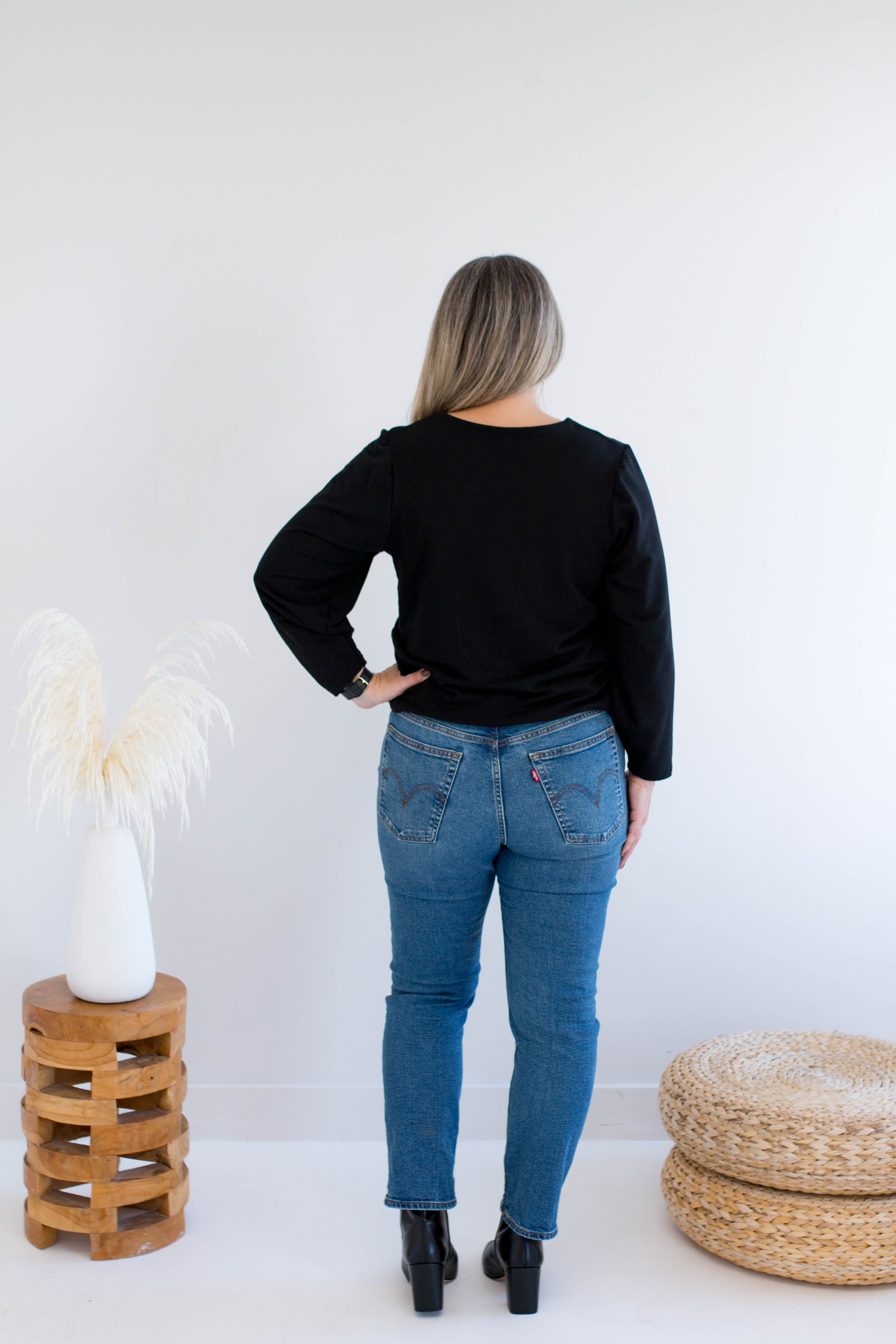 back view of a woman wearing a black v-neck sweater and blue jeans standing against a white wall with decorative items.