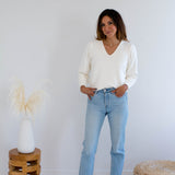 Woman wearing a cream v-neck  sweater and light blue jeans standing in a minimalistic room with a wooden stool and woven basket