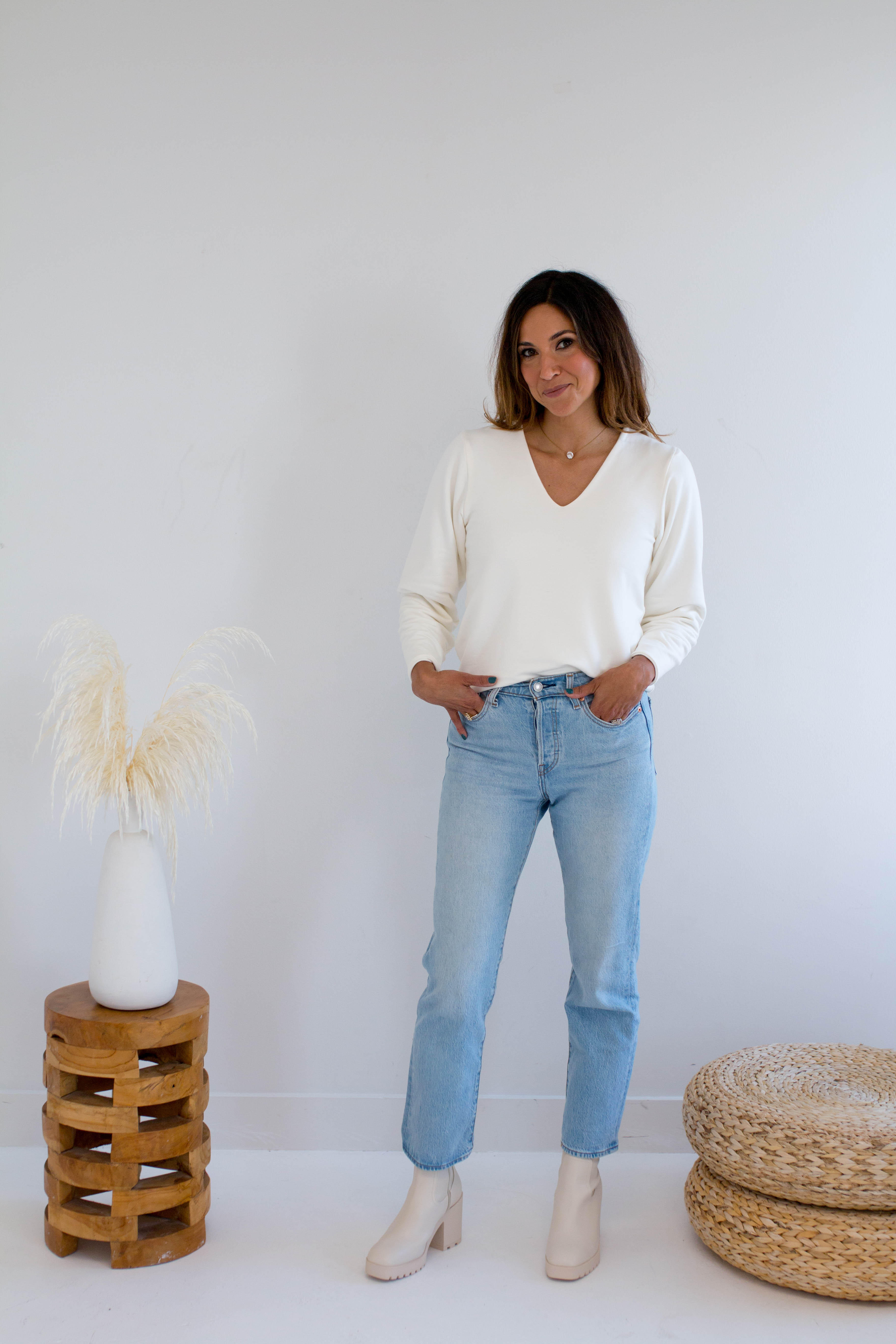 Woman wearing a cream v-neck  sweater and light blue jeans standing in a minimalistic room with a wooden stool and woven basket
